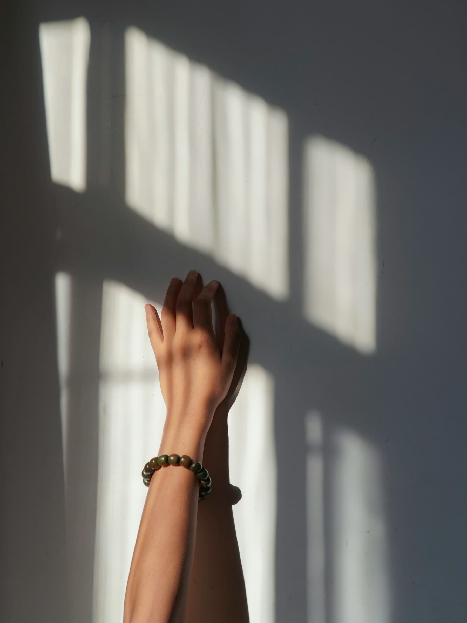 A raised forearm and hand against a grey wall striped with window light, a thin bracelet at the wrist.