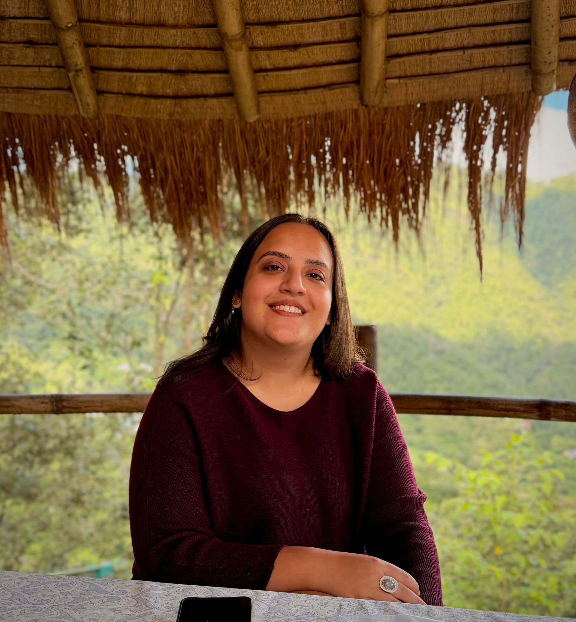 Laksheta Moorjani smiling, seated at a table under a thatched roof with green hills behind her.