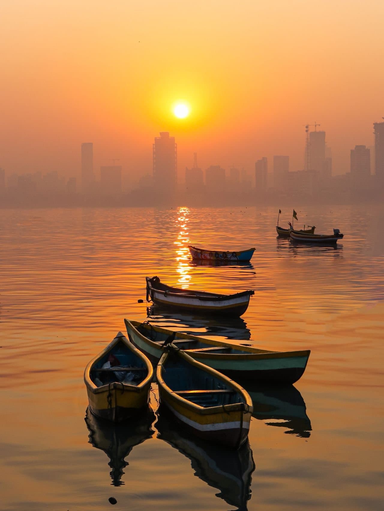 Small wooden fishing boats resting on still water at sunrise, the Mumbai skyline soft in the haze behind them.