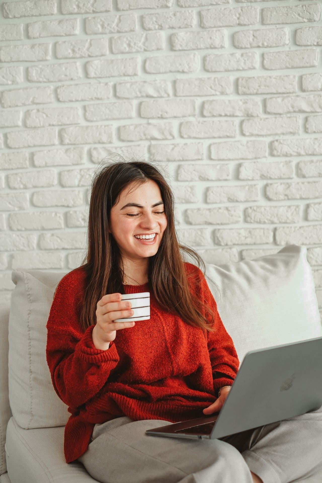 A woman in a red jumper laughing on a sofa at home, a cup in one hand and a laptop on her lap.