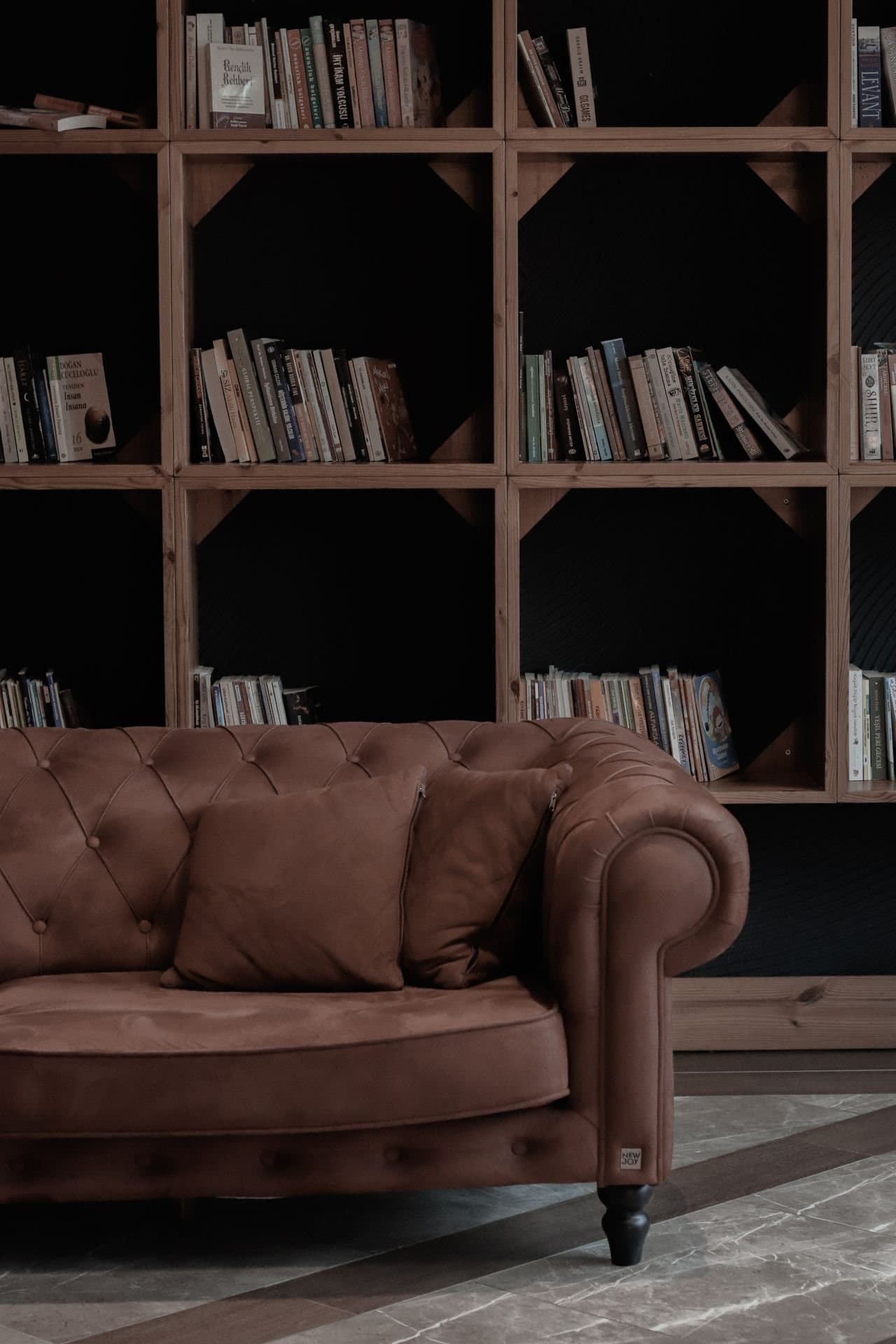 A worn tan leather sofa with two cushions in front of wooden shelves of books.